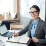 Professionnelle souriante en bureau, prend des notes dans un carnet près d’un ordinateur; pose d'un planning pour comment bien préparer son déménagement.