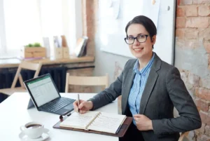 Professionnelle souriante en bureau, prend des notes dans un carnet près d’un ordinateur; pose d'un planning pour comment bien préparer son déménagement.